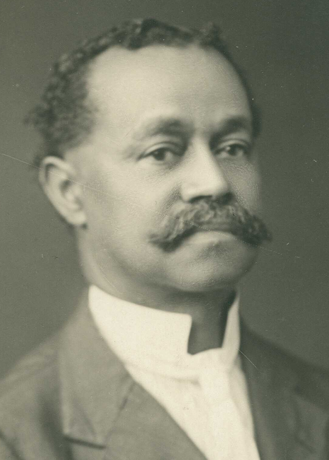 An older photograph shows a man with a mustache wearing a suit and high-collared shirt. He has short, curly hair and is facing the camera with a neutral expression, in front of a plain background.