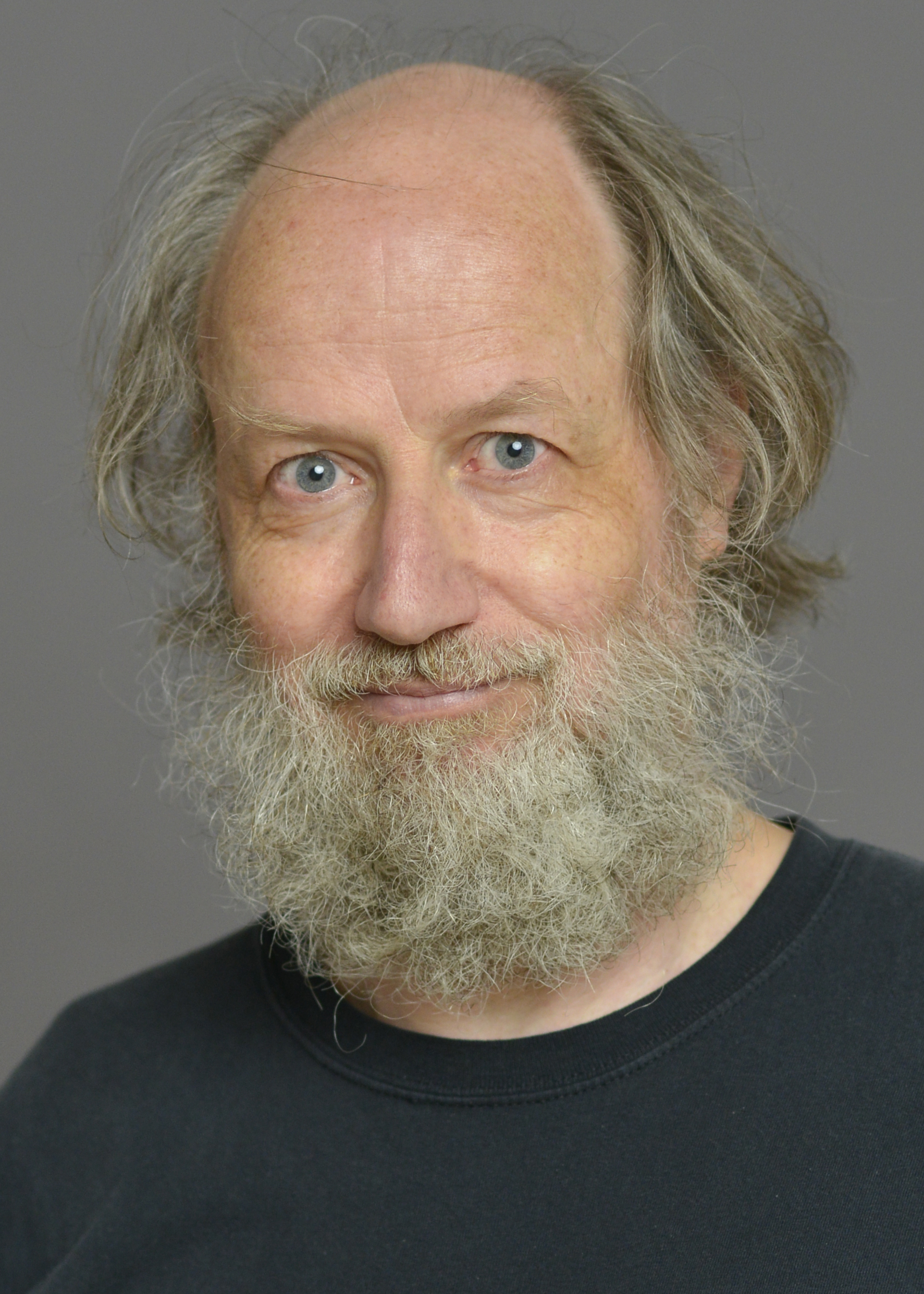 A man with a balding head and a full, graying beard is shown against a plain background. He is wearing a black shirt and has blue eyes.