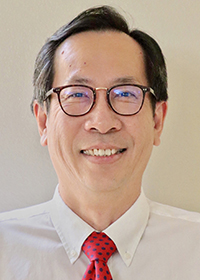 A man with glasses and short dark hair smiles at the camera. He is wearing a white shirt with a red tie that has small blue spots. The background is a plain light-colored wall.