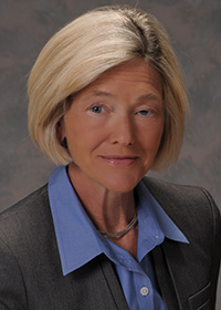 A woman with chin-length blonde hair is wearing a business suit with a grey jacket and a blue collared shirt. She is posing against a grey background, looking straight at the camera with a slight smile.