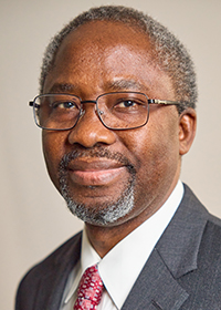 A man with short gray hair, a beard, and glasses wears a dark suit, white shirt, and red patterned tie. He is looking into the camera and smiling slightly. The background is plain and gray.