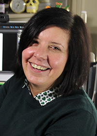 A woman with shoulder-length dark hair is smiling at the camera. She is wearing a dark green sweater over a collared shirt with a pattern of small black insects and green hearts. The background includes computer screens and various office items.