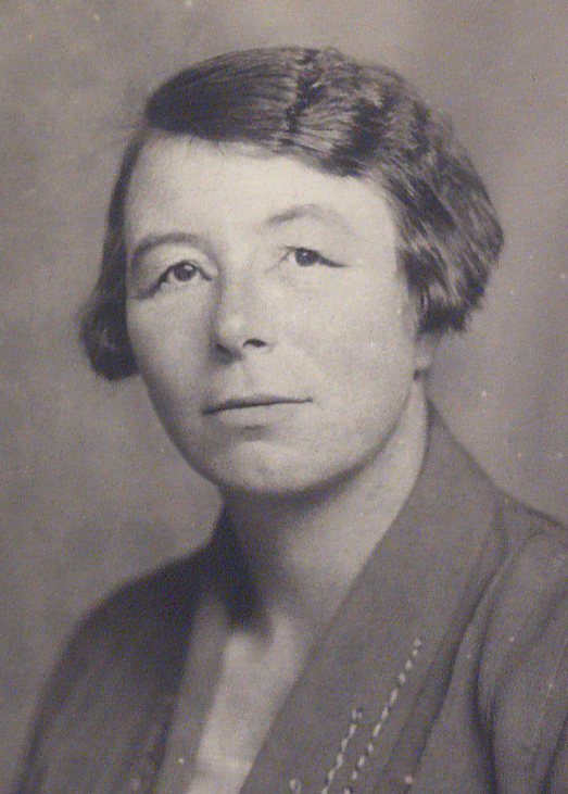 Black and white portrait of a woman with short, wavy dark hair, wearing a collared garment. She faces the camera with a neutral expression against a plain background.