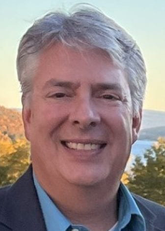 A man with gray hair is smiling at the camera. He is wearing a blue collared shirt and a dark jacket. The background shows trees with fall foliage and a clear sky.