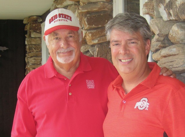 Two men stand close together and smile at the camera in front of a stone wall. Both wear red Ohio State shirts; the older man on the left also wears an Ohio State Buckeyes hat.