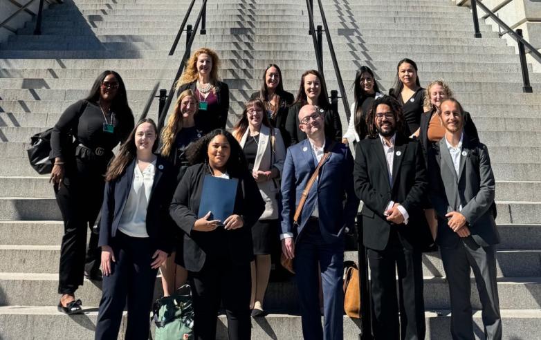 A group of people dressed in formal attire stand on a set of outdoor stone steps. They are posing for a group photo, and many are holding folders or bags. The background features metal railings along the steps.