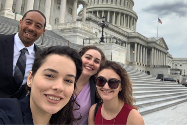 Four people, part of the Science Policy Fellows program, stand smiling before the U.S. Capitol, where they learn to advocate for entomologists and federal policy in Washington, D.C.; columns, steps, and an American flag are seen in the background.