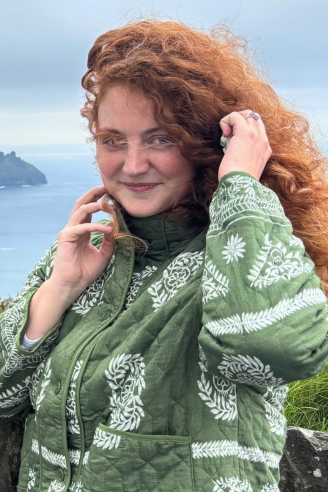A woman with curly red hair wearing a green patterned jacket stands outdoors by the sea, holding their hair with one hand.